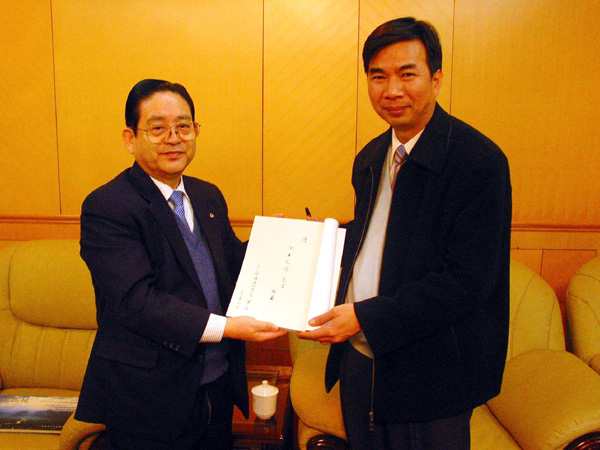 梁宏校长请李刚寿先生（左）将其亲笔签名的书转赠予池田大作先生。